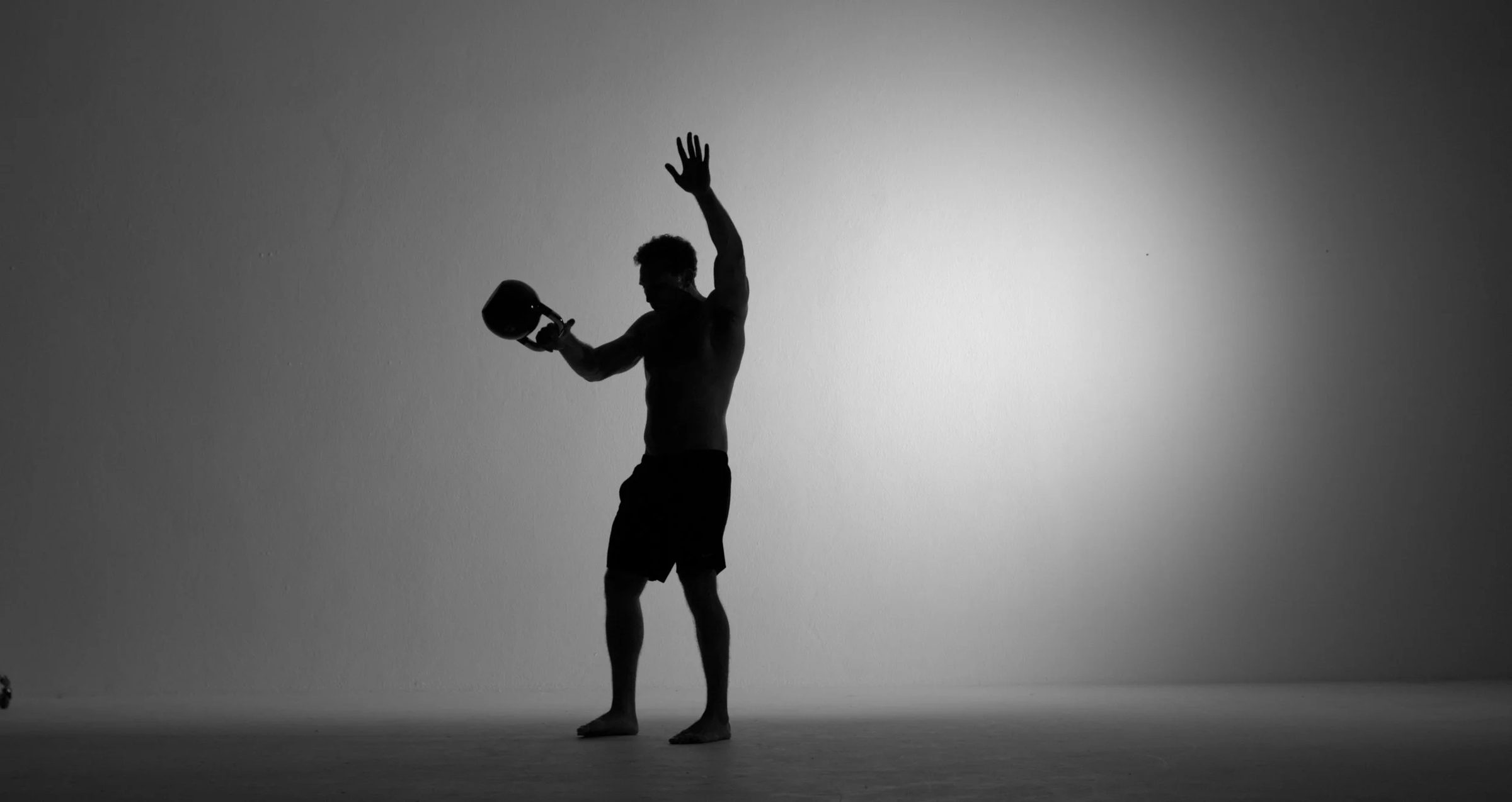 Praticante a elevar um kettlebell acima da cabeça, em silhueta contra a parede do estúdio